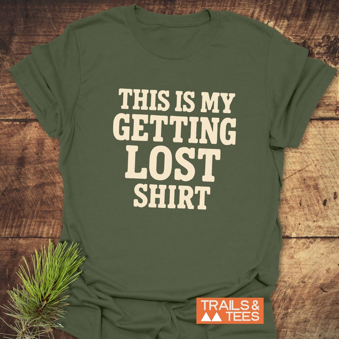 This Is My Getting Lost T-Shirt in cream block letters on olive-green fabric, with an orange TRAILS & TEES logo; laid on a wooden surface with pine needles.