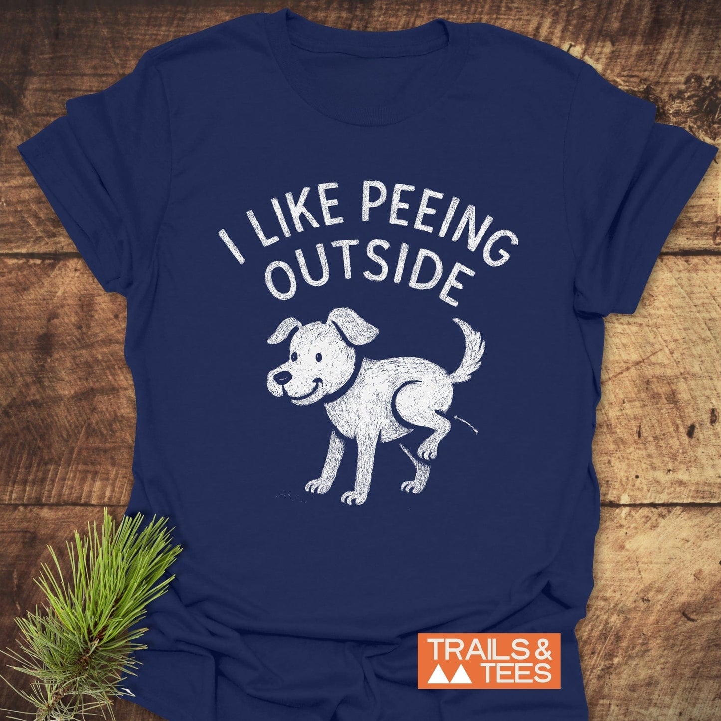 I Like Peeing Outside T-Shirt features a white graphic of a dog on a navy-blue tee, styled with an evergreen sprig, highlighting outdoor adventure and comfort.