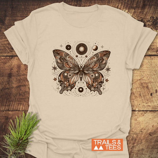 Celestial Butterfly T-Shirt featuring a stylized butterfly with celestial motifs, laid on wood with TRAILS & TEES logo; showcases adventure-ready, sustainable design.