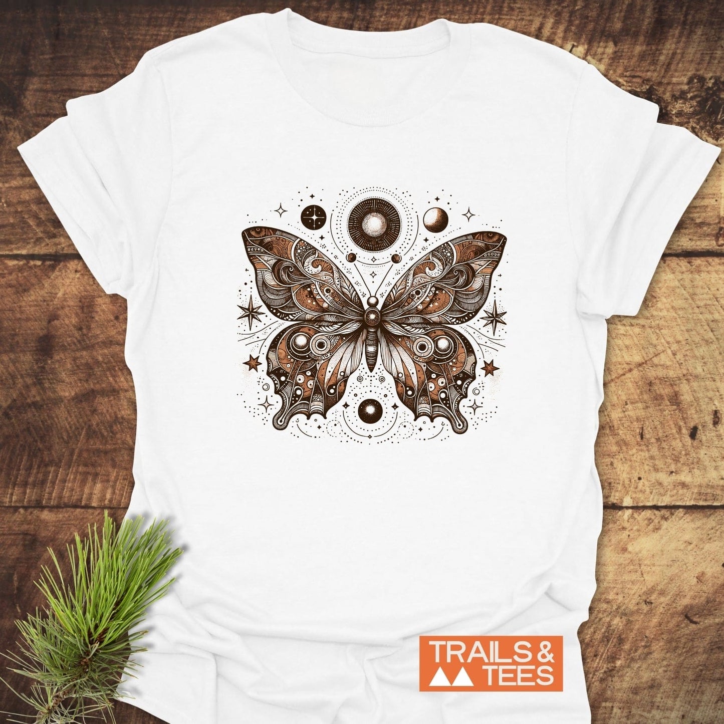 Celestial Butterfly T-Shirt featuring a detailed butterfly with celestial motifs on soft cotton, laid on wood beside a pine sprig, with TRAILS & TEES logo.