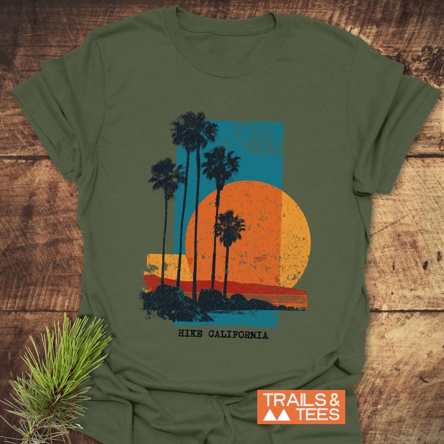 Hike California T-Shirt with a retro sun and palm graphic, laid on wood. Features soft cotton, durable print, and typewriter-style text.
