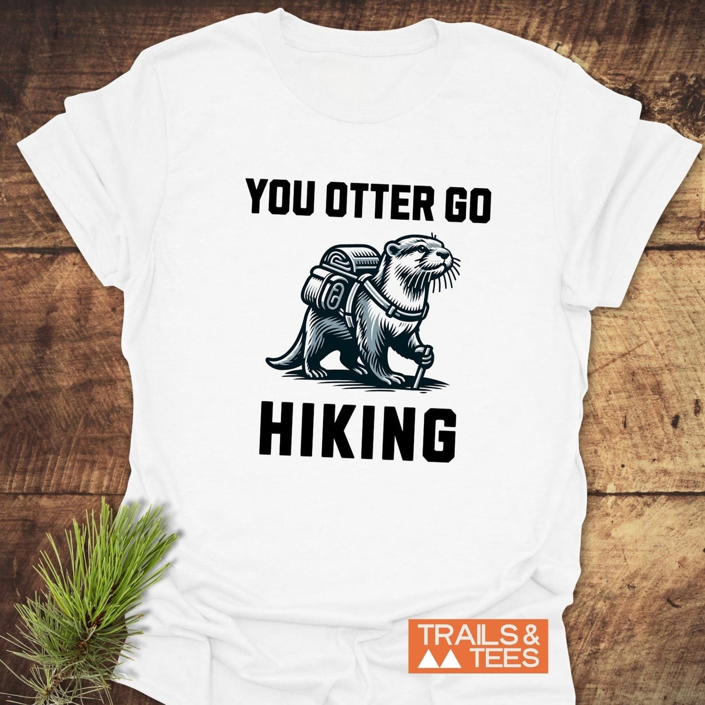 You Otter Go Hiking T-Shirt: white with line-art otter print, slogan, and Trails & Tees logo. Laid on wood with pine sprig for outdoorsy feel.