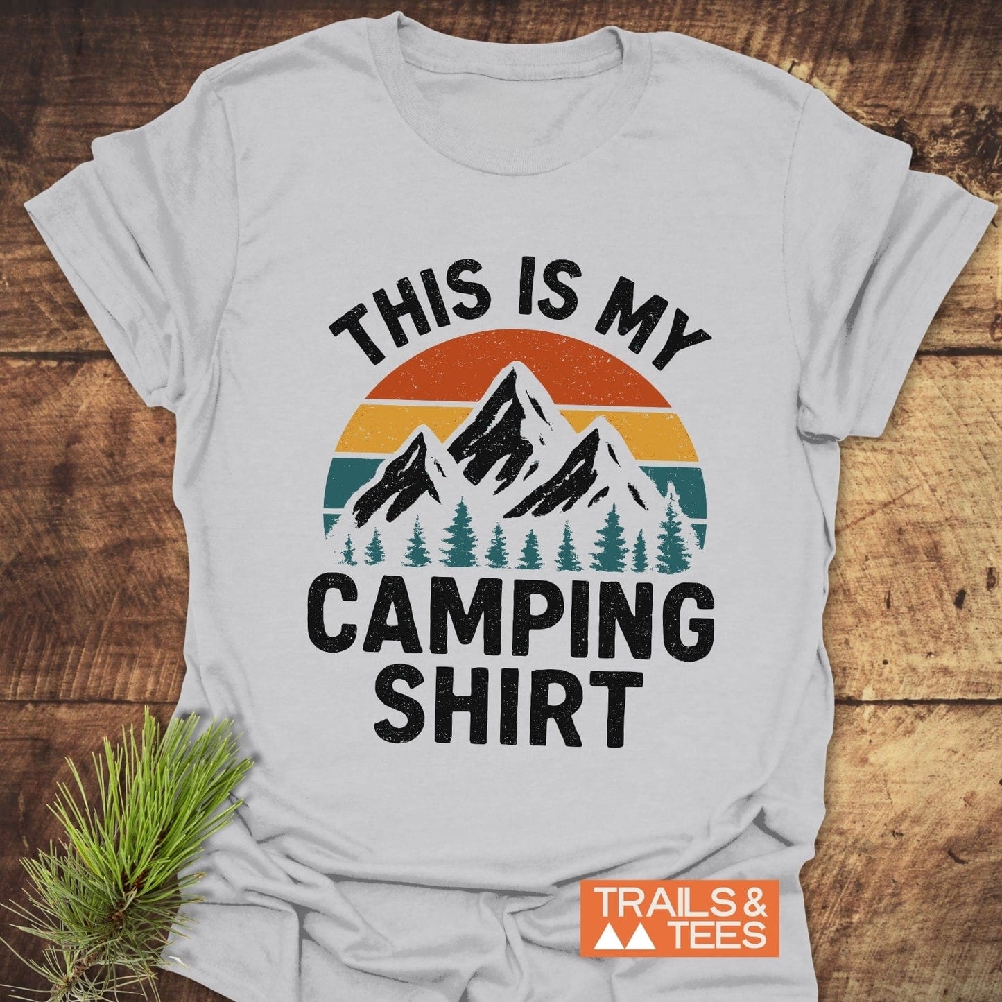 This Is My Camping T-Shirt, featuring a sunset and mountain graphic, laid on wood with a pine sprig, embodies outdoor adventure and comfort.
