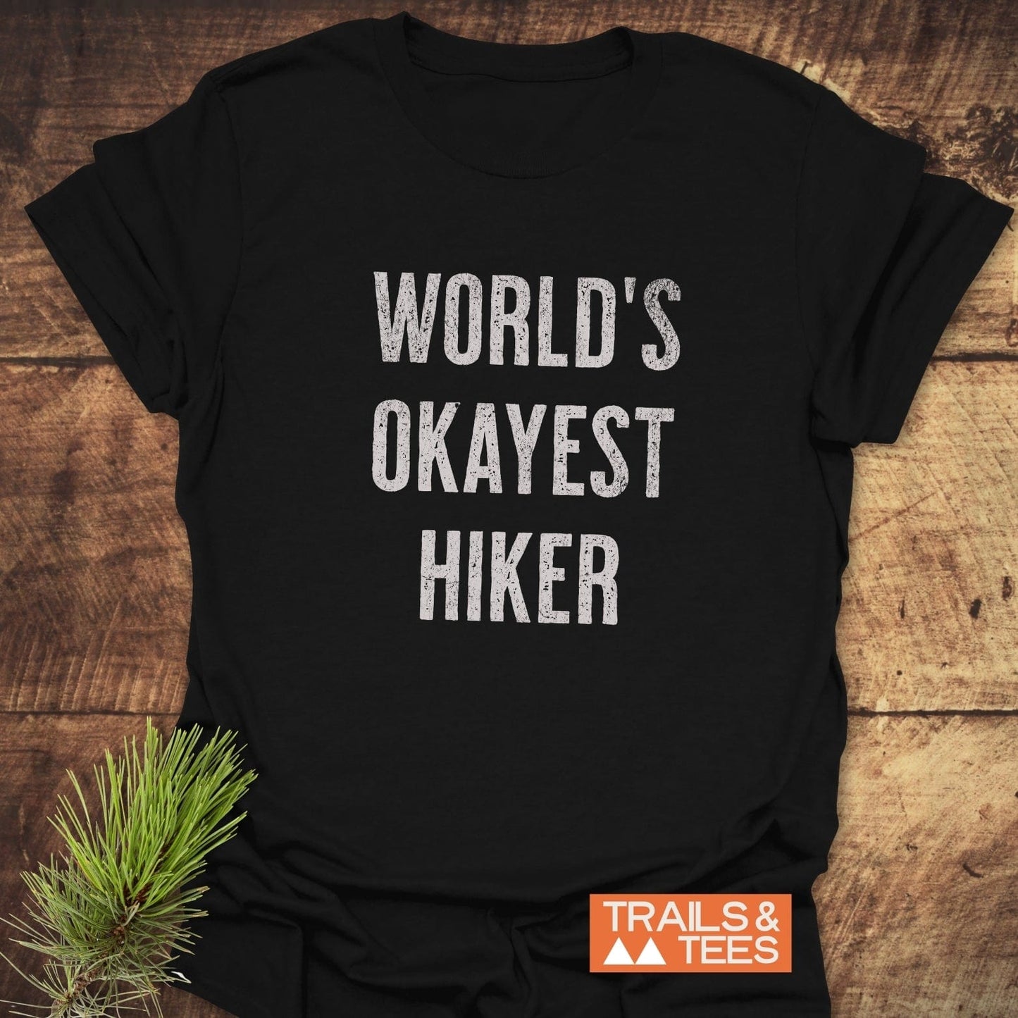 Black T-shirt with WORLD’S OKAYEST HIKER slogan, TRAILS & TEES logo, and pine sprig; crafted for comfort with durable, pre-shrunk cotton.