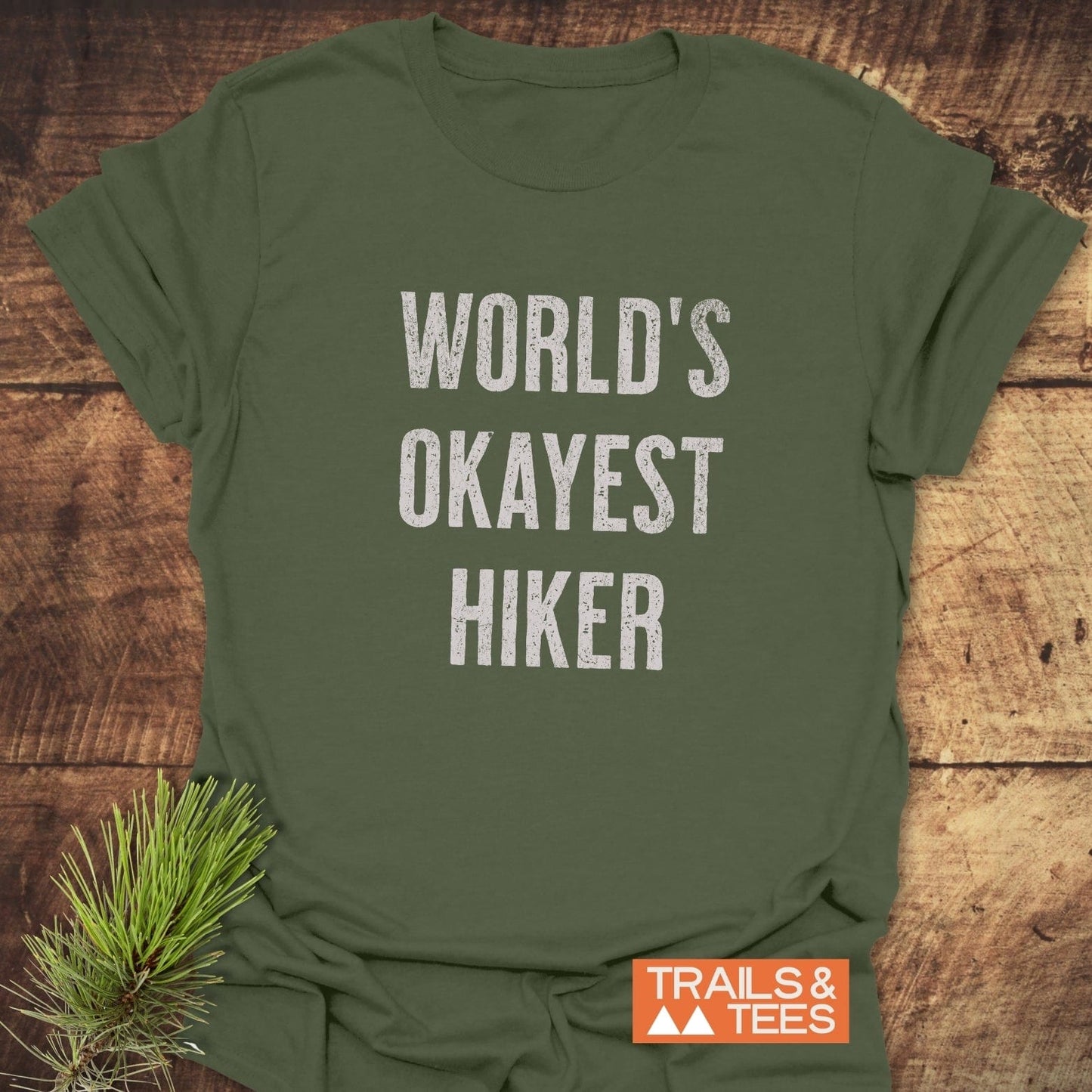 Olive-green World's Okayest Hiker T-shirt on rustic wood, featuring distressed white lettering. Includes a TRAILS & TEES logo, highlighting outdoor adventure spirit.