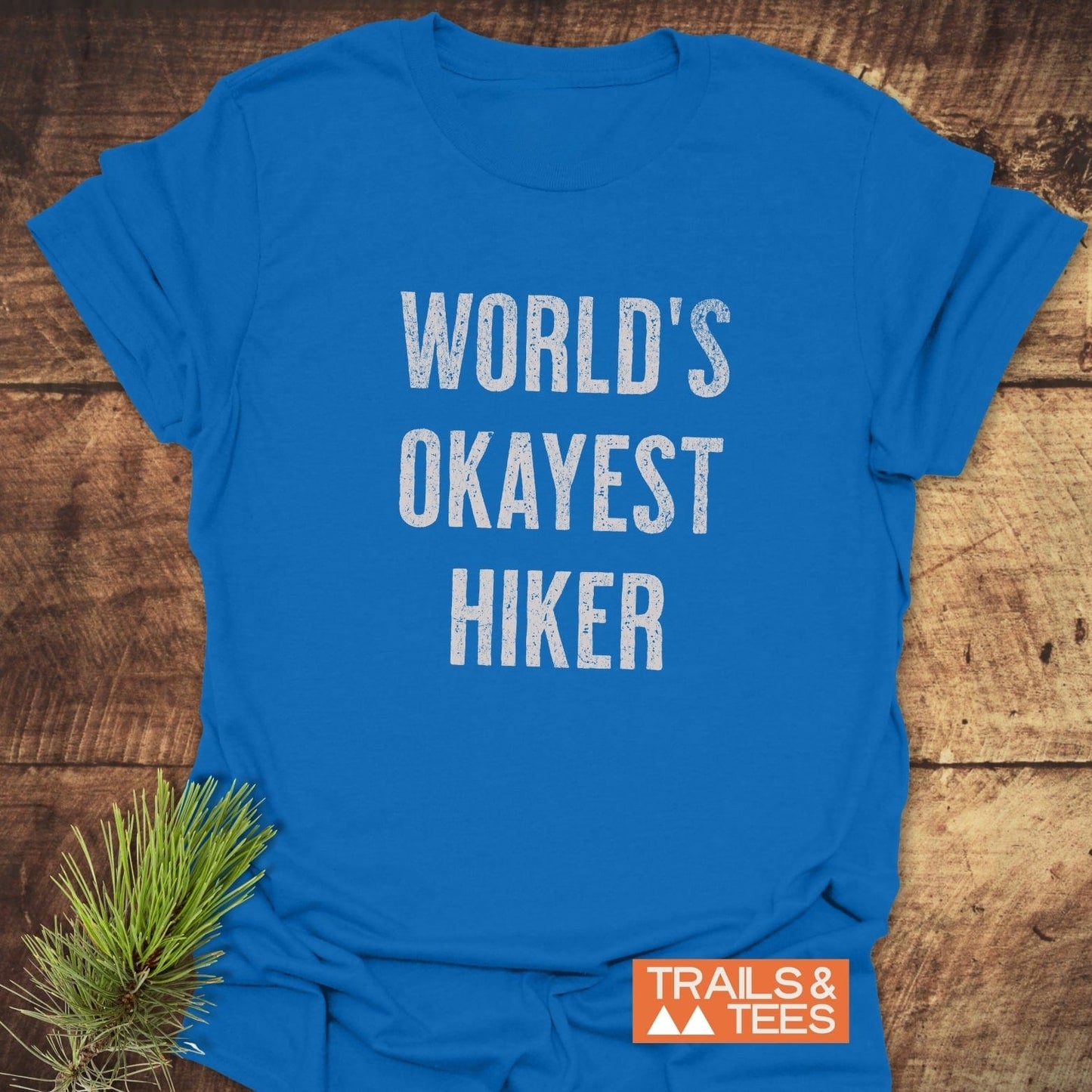 World's Okayest Hiker T-Shirt in bright blue with distressed white lettering, showcasing outdoor theme. Includes Trails & Tees logo, pine sprig for rustic touch.
