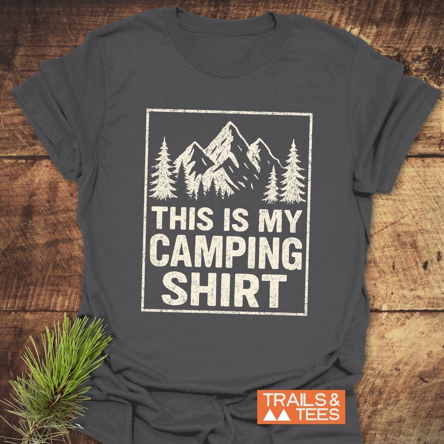 This Is My CAMPING T-Shirt featuring mountain graphic and text, laid flat with TRAILS & TEES label; soft, durable ring-spun cotton, perfect for outdoor enthusiasts.