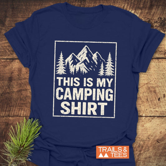 Navy-blue This Is My CAMPING T-Shirt with white print of pine trees and mountains, laid on wood with a pine sprig. Features include soft cotton and durable print.