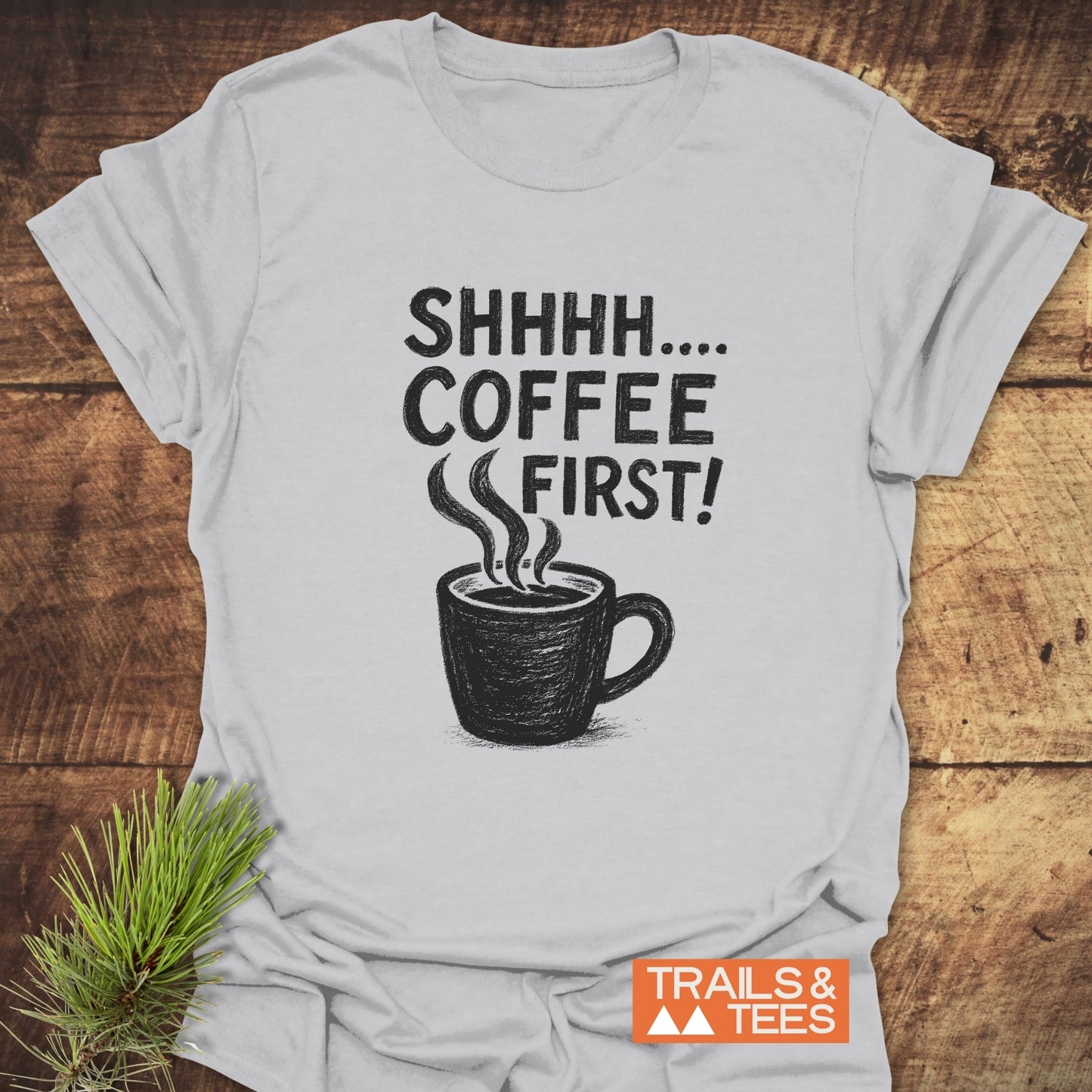 Shhhhh, Coffee First T-Shirt with a black coffee mug graphic and bold lettering, crafted from soft ring-spun cotton, laid flat on a wooden surface.