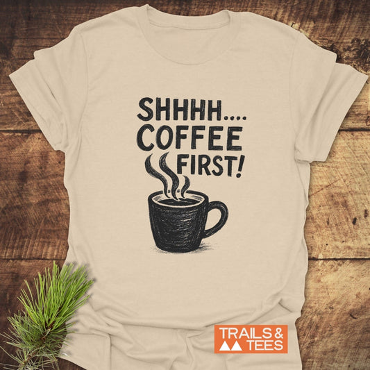 Light-beige Shhhhh, Coffee First T-Shirt featuring a coffee cup illustration and text, laid on wood. Includes TRAILS & TEES logo, emphasizing comfort and durability.