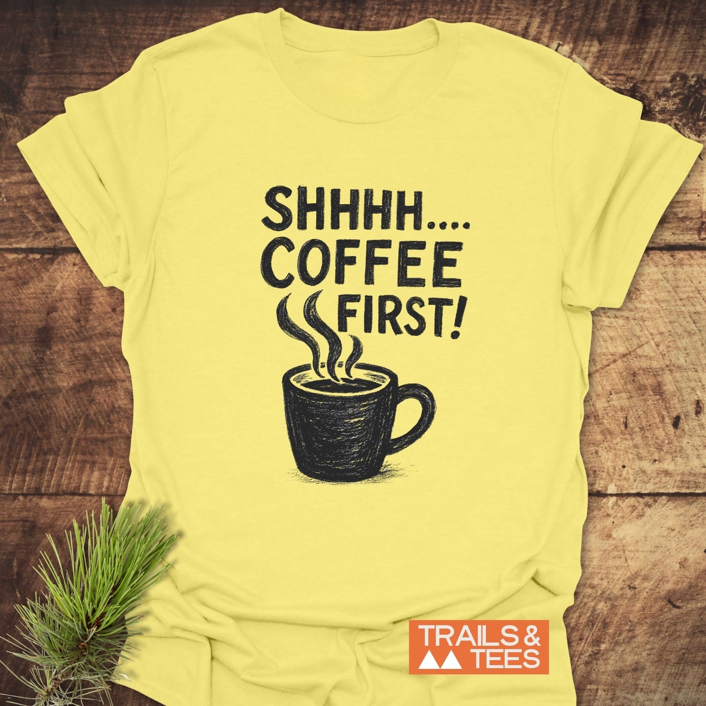 Shhhhh, Coffee First T-Shirt with a black coffee mug illustration and slogan, laid flat with pine sprig, showcasing durable comfort and outdoor-inspired design.