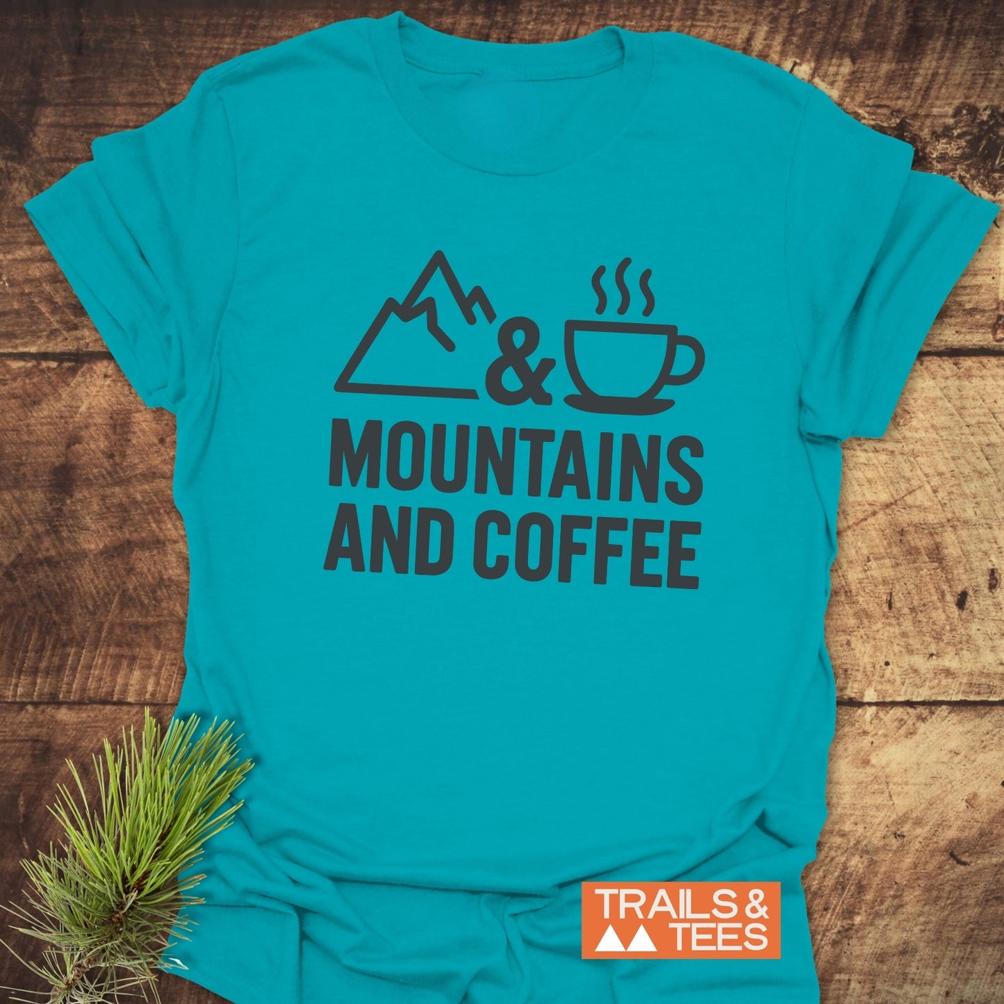 Mountains And Coffee T-Shirt featuring mountain icon, ampersand, coffee cup, and text, laid on wood with pine sprig and TRAILS & TEES label.