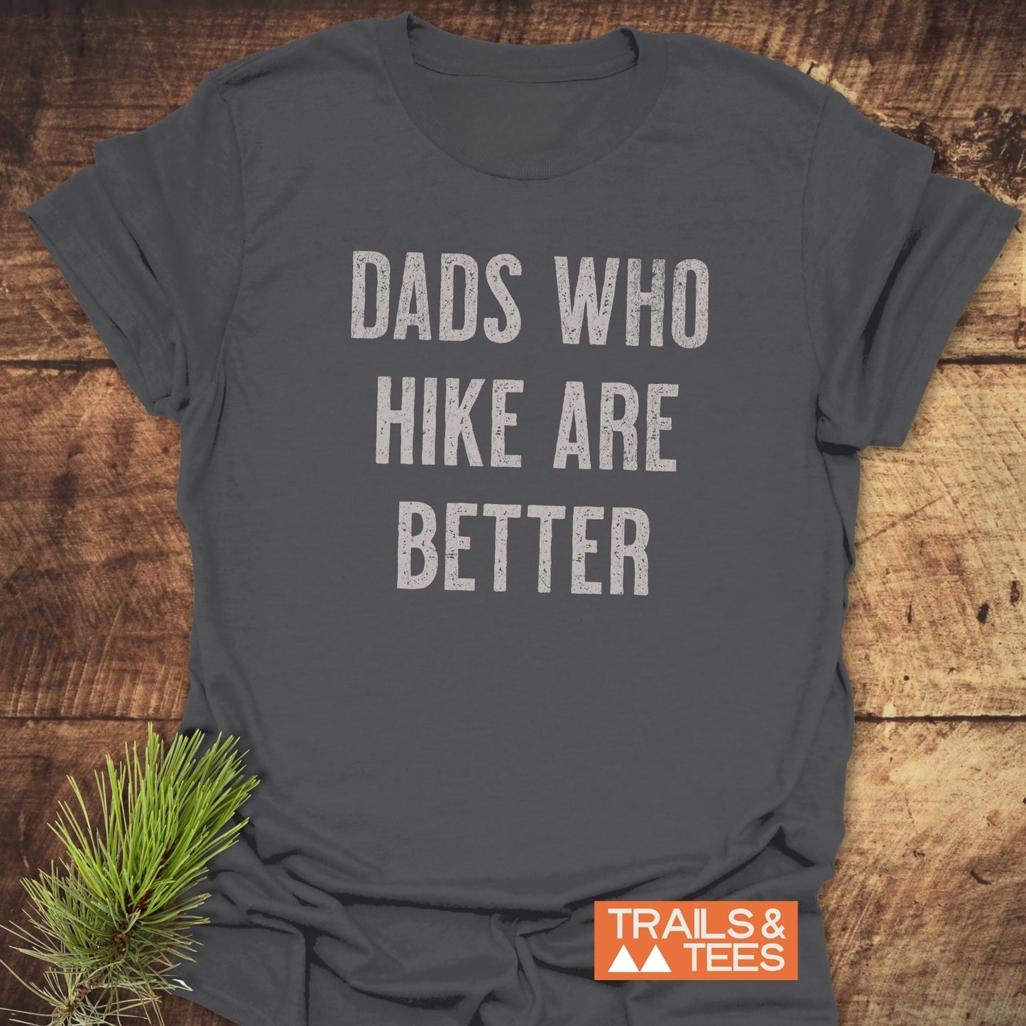 Dark-gray Dads Who Hike Are Better T-shirt on wood, featuring distressed light lettering, small orange Trails & Tees logo, and pine sprig accent.