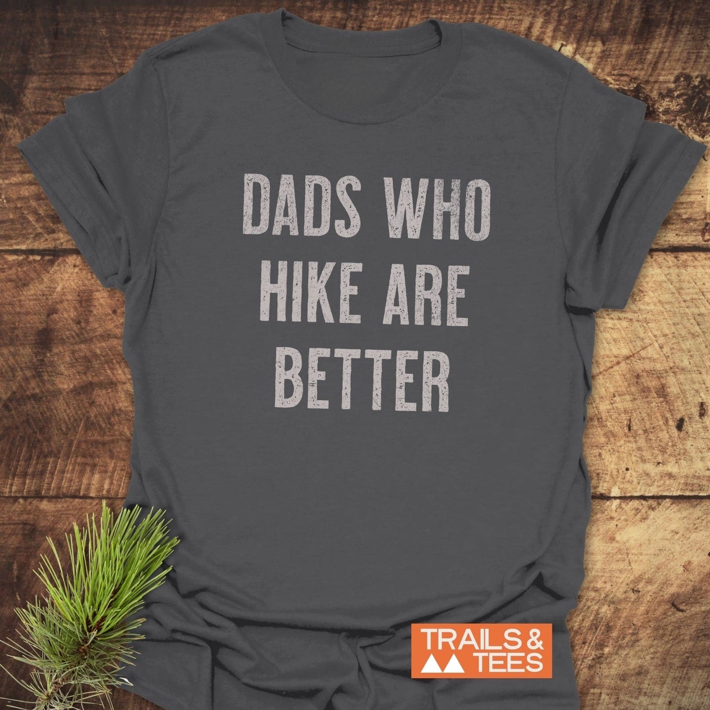 Dark-gray Dads Who Hike Are Better T-shirt on wood, featuring distressed light lettering, small orange Trails & Tees logo, and pine sprig accent.