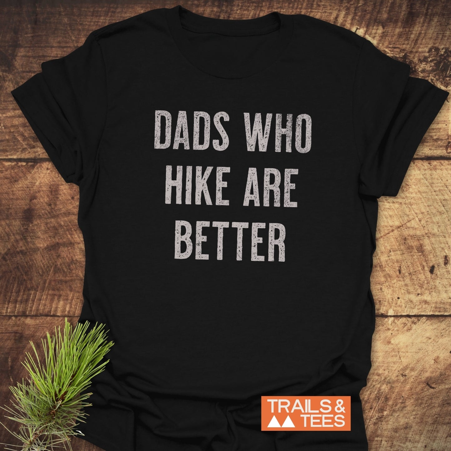 Black T-shirt with DADS WHO HIKE ARE BETTER in white distressed lettering, featuring TRAILS & TEES logo, laid on wood, emphasizing outdoor adventure spirit.