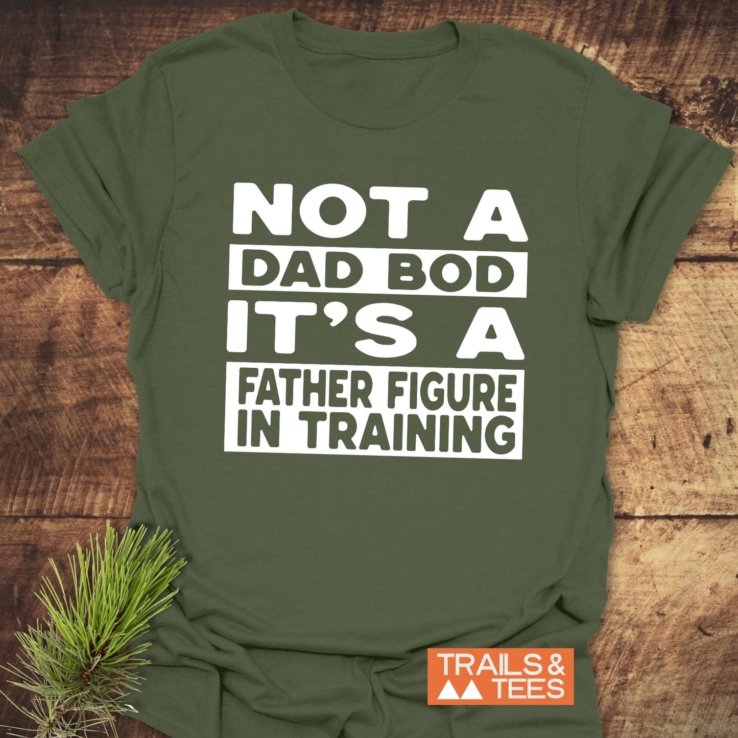 It's Not A Dad Bod T-Shirt featuring bold white text, laid on wood with pine sprig, Trails & Tees logo visible. Crafted for outdoor comfort and style.