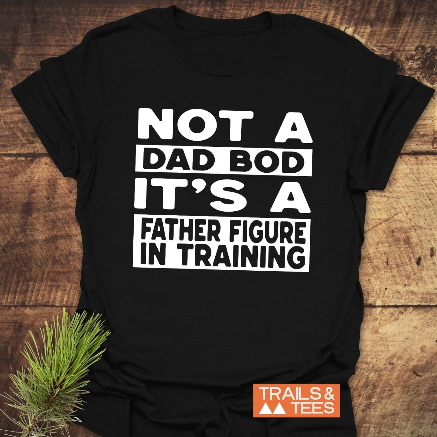 It's Not A Dad Bod T-Shirt with bold slogan, laid flat, features soft ring-spun cotton, durable print, and Trails & Tees logo beside a pine sprig prop.