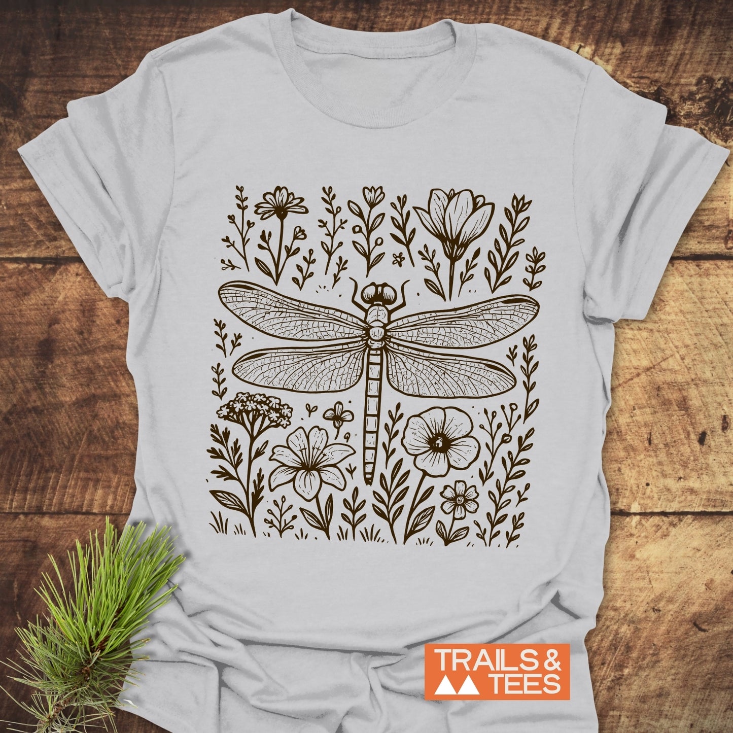 Dragonfly T-Shirt featuring a detailed dragonfly line drawing with wildflowers, laid on wood. Includes “TRAILS & TEES” logo and pine branch accent.