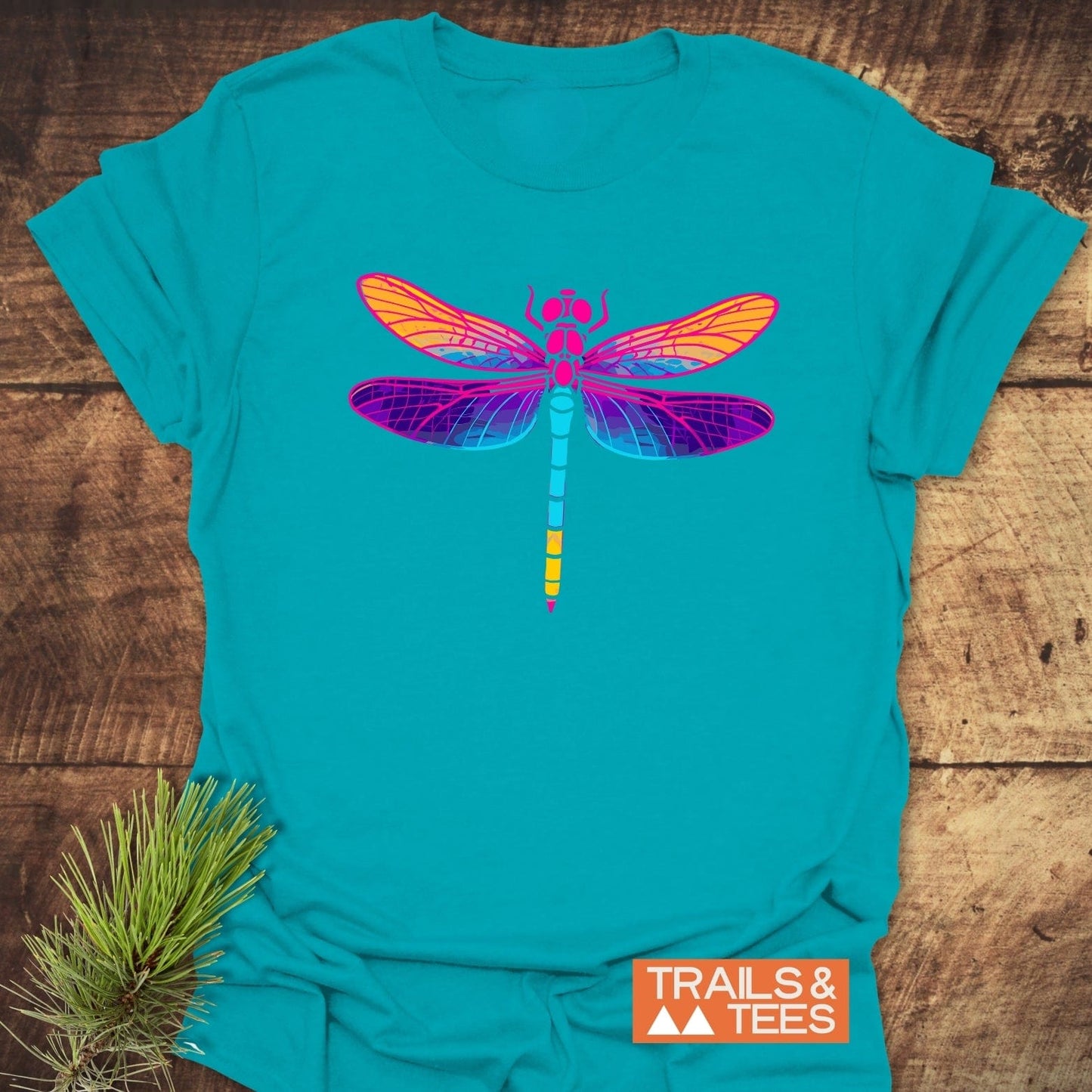 Psychedelic Dragonfly T-Shirt in teal, featuring a vibrant dragonfly graphic, on rustic wood. Includes TRAILS & TEES logo, emphasizing outdoor-inspired design.
