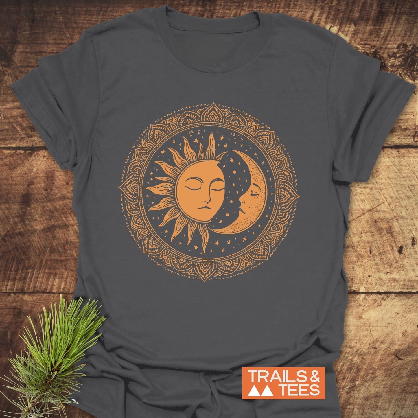 Bohemian Sun And Moon T-Shirt featuring a burnt-orange celestial design on charcoal fabric, laid on wood with pine sprig, showcasing durable, eco-friendly craftsmanship.