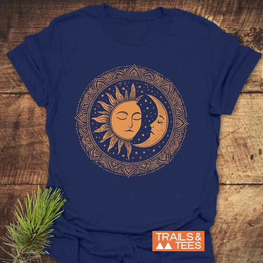 Bohemian Sun And Moon T-Shirt featuring a golden-orange mandala design with sun and moon motifs, laid on a wooden surface with a pine sprig.