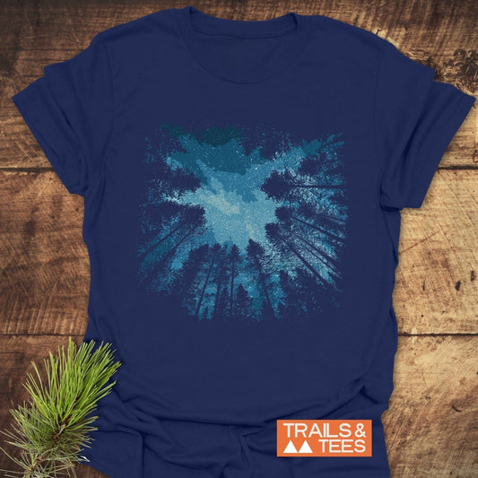 Starry Night Forest T-Shirt with pine tree and starry sky illustration, navy blue, featuring TRAILS & TEES logo, resting on wooden surface.