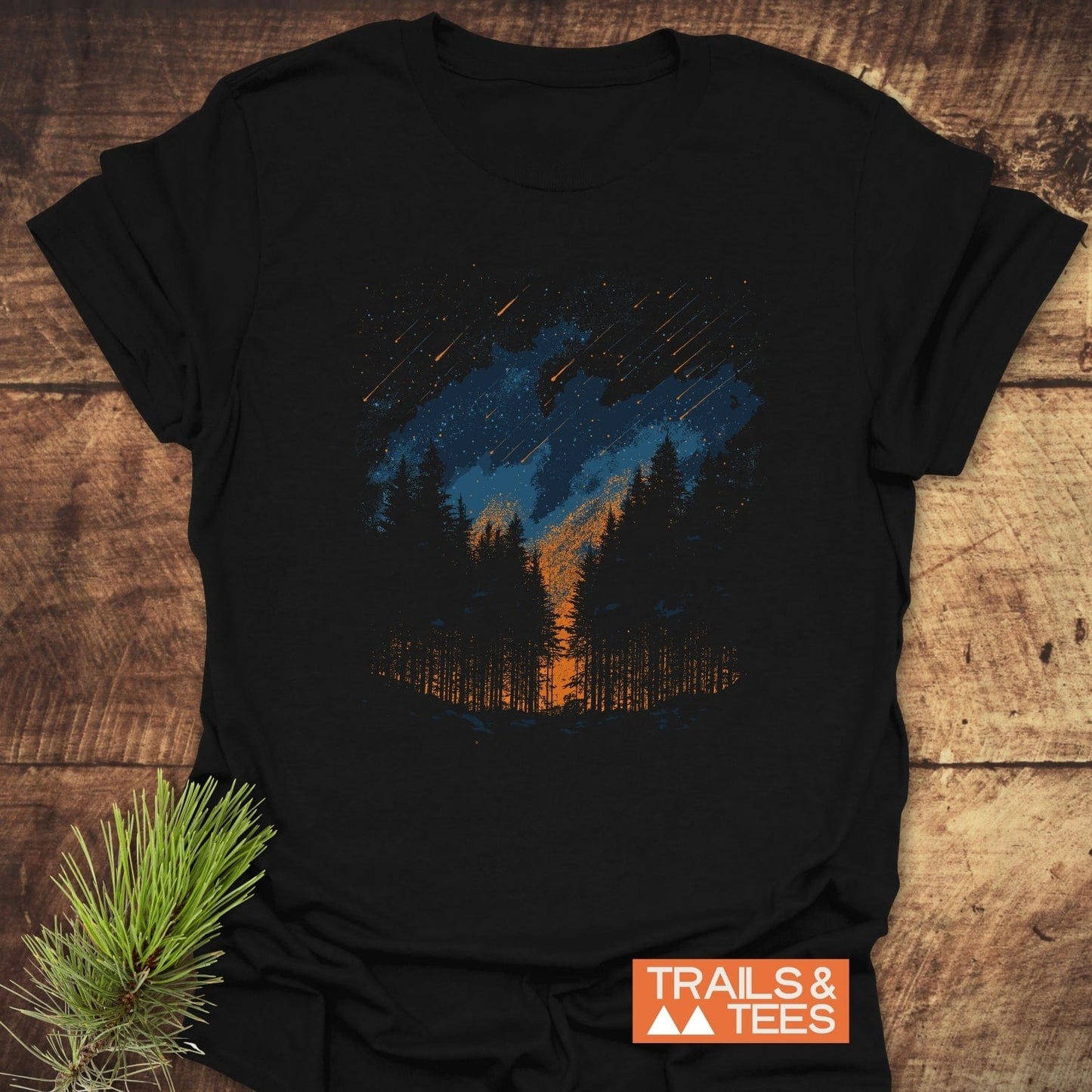 Sunset Forest T-Shirt featuring a stylized forest scene with pine trees, orange horizon, and shooting stars, laid flat with Trails & Tees logo visible.