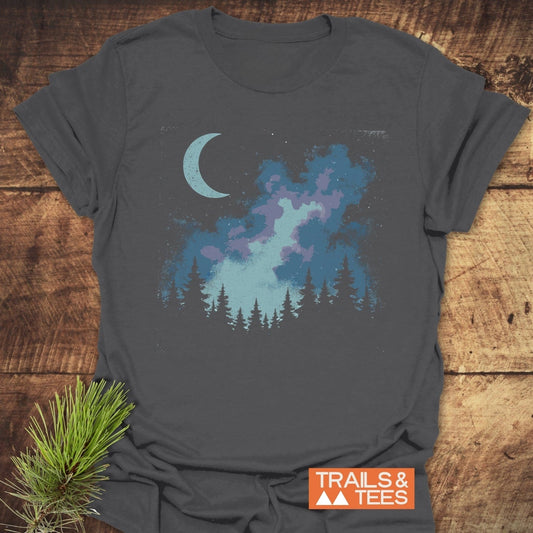 Moonlit Forest T-Shirt on wooden surface, featuring a night-scene graphic with a crescent moon, nebula, and silhouetted pine trees; overlaid TRAILS & TEES logo.