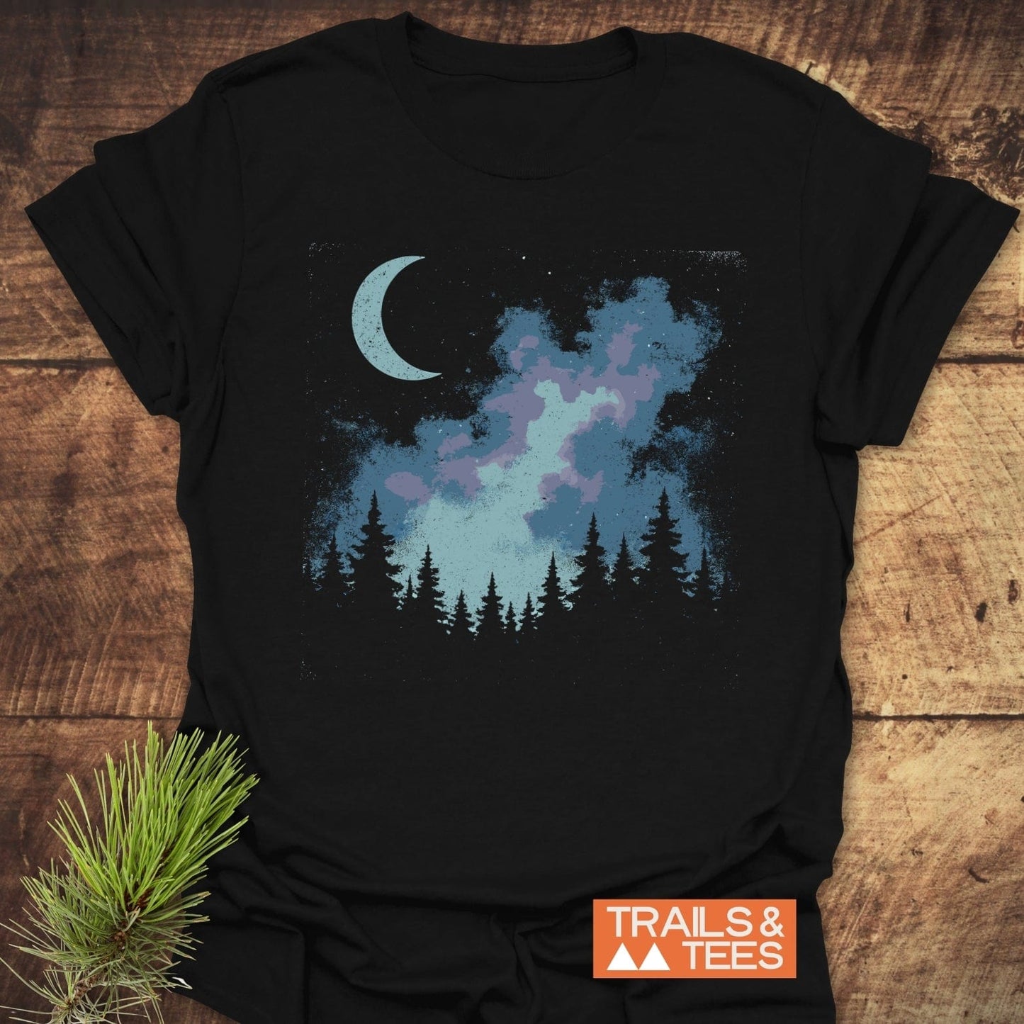 Moonlit Forest T-Shirt featuring a night scene with a crescent moon, colorful nebula, silhouetted pines, and the TRAILS & TEES logo, laid on wood.