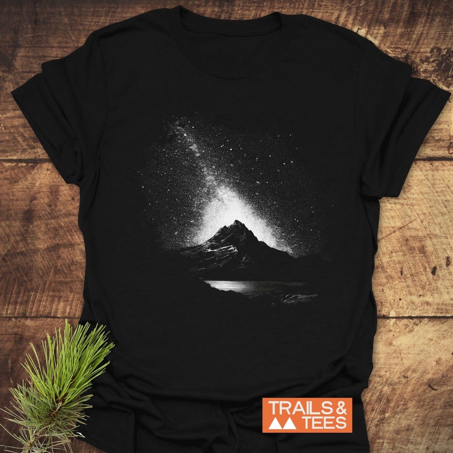 Starry Sky Above T-Shirt with mountain and Milky Way graphic, laid on wood, featuring TRIALS & TEES logo, soft cotton, pre-shrunk, durable print.