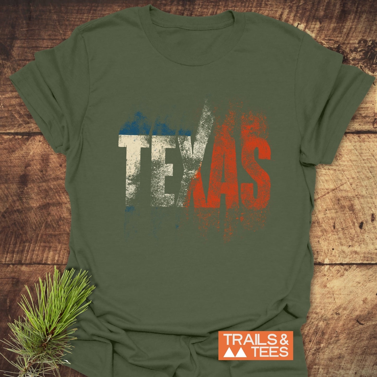 Olive-green TEXAS T-Shirt by TRAILS & TEES, featuring Texas-flag colors in distressed print, laid flat on rustic wood with pine sprig accent.