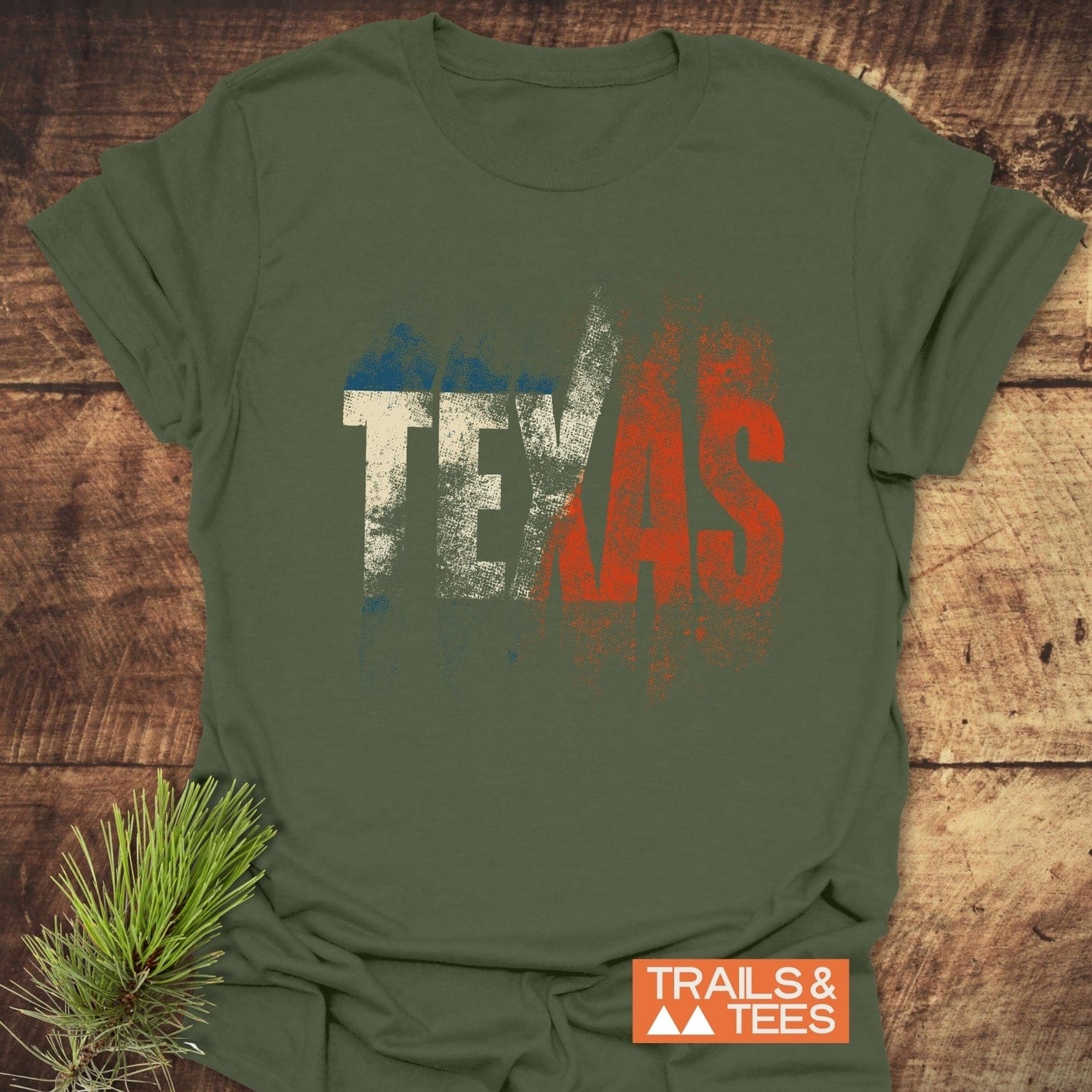 Olive-green TEXAS T-Shirt by TRAILS & TEES, featuring Texas-flag colors in distressed print, laid flat on rustic wood with pine sprig accent.