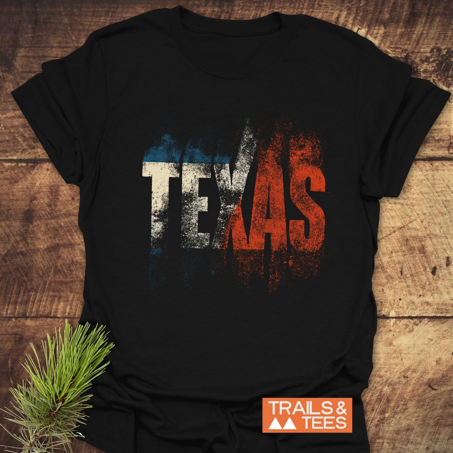 Black TEXAS T-Shirt with distressed lettering in white and red on soft, lightweight cotton, styled with pine sprigs. Featuring Trails & Tees logo.