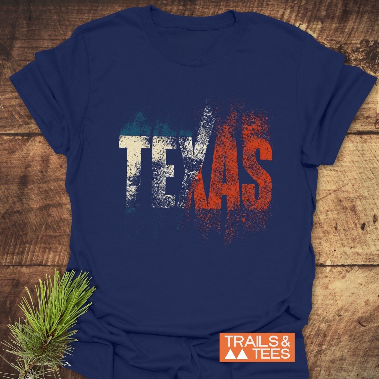 Navy-blue TEXAS T-Shirt with distressed flag colors, laid on wood. Features TRAILS & TEES logo, pine branch accent. Soft, durable, and pre-shrunk.