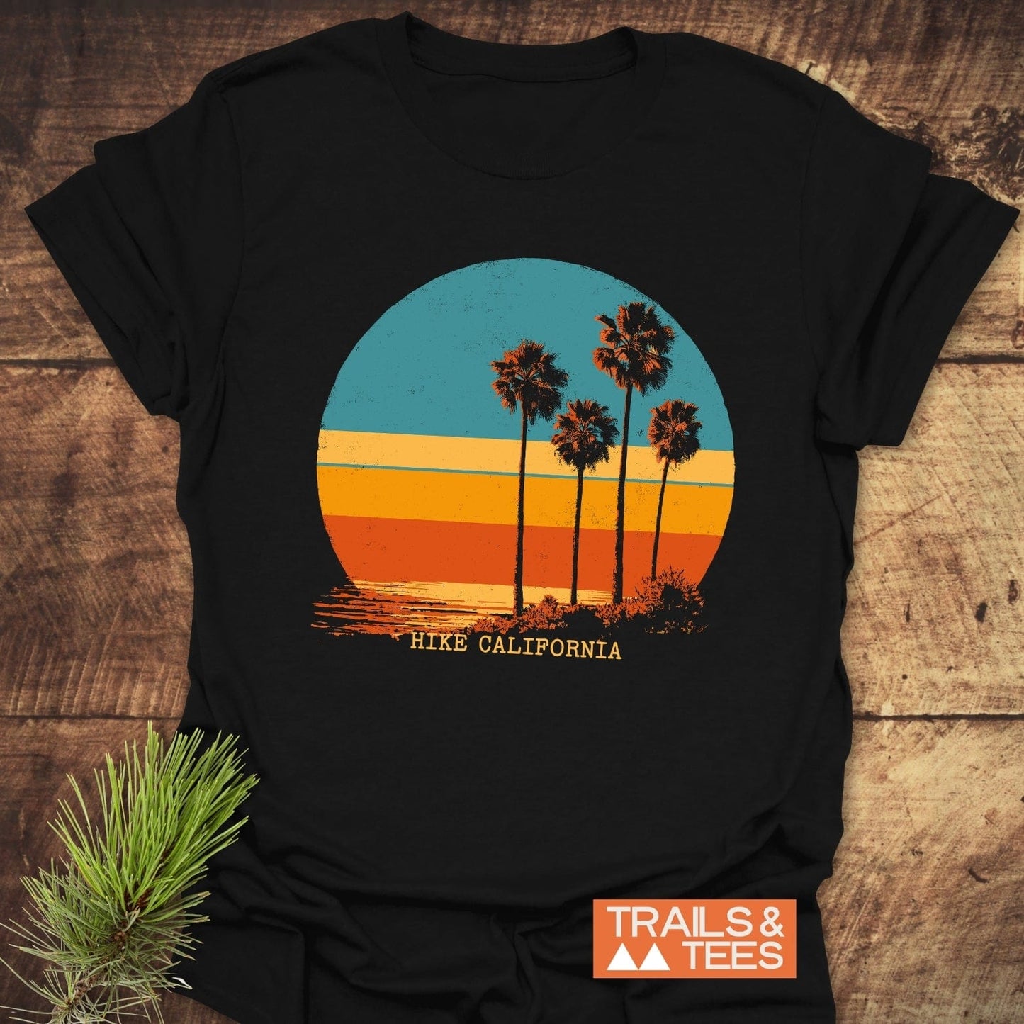 Hike California T-Shirt on wood, features a vintage sunset with palm trees, HIKE CALIFORNIA text, and TRAILS & TEES logo, pine sprig beside.