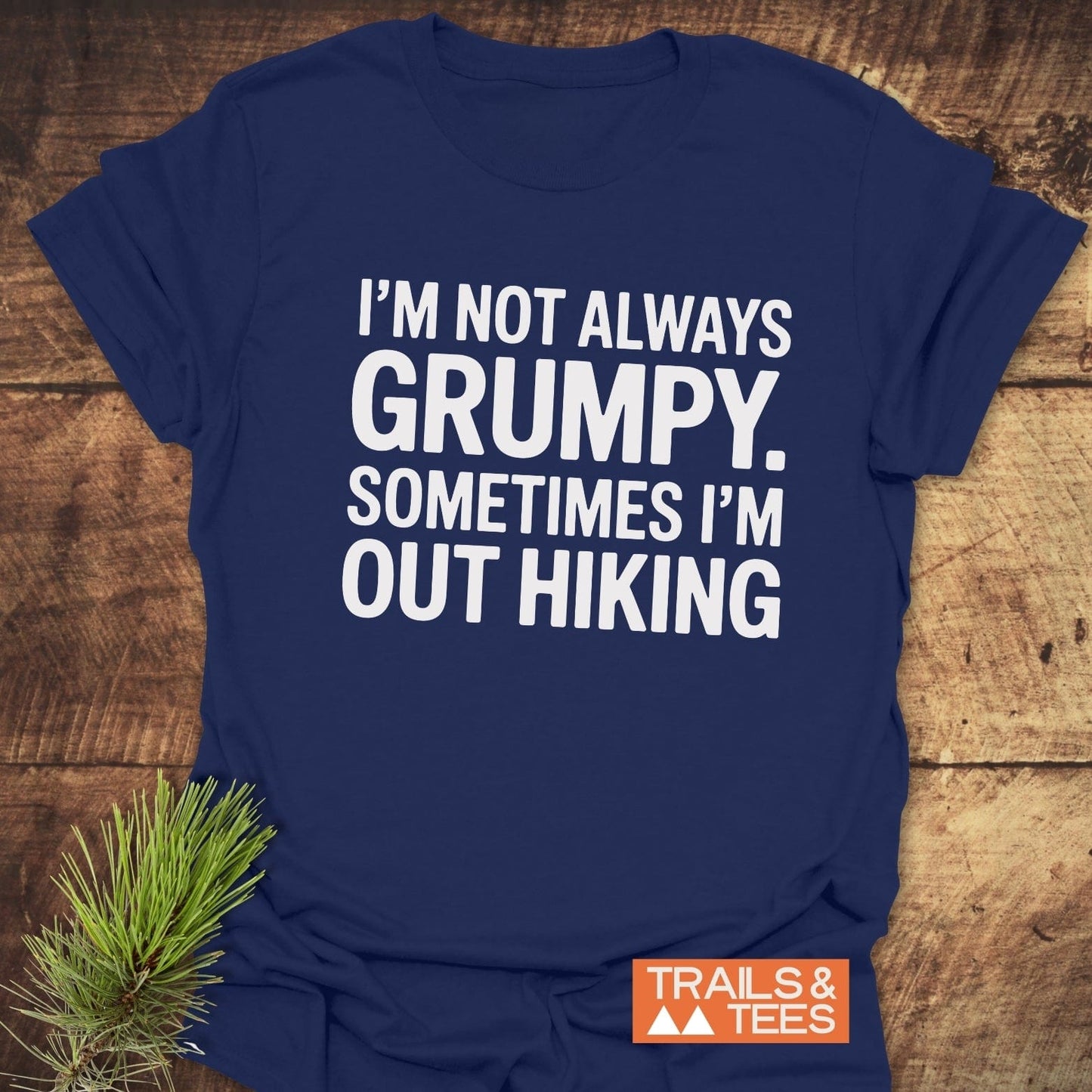 Im Not Always Grumpy T-Shirt on wooden surface, featuring bold white text and Trails & Tees logo. Pine sprig beside it enhances the outdoor theme.