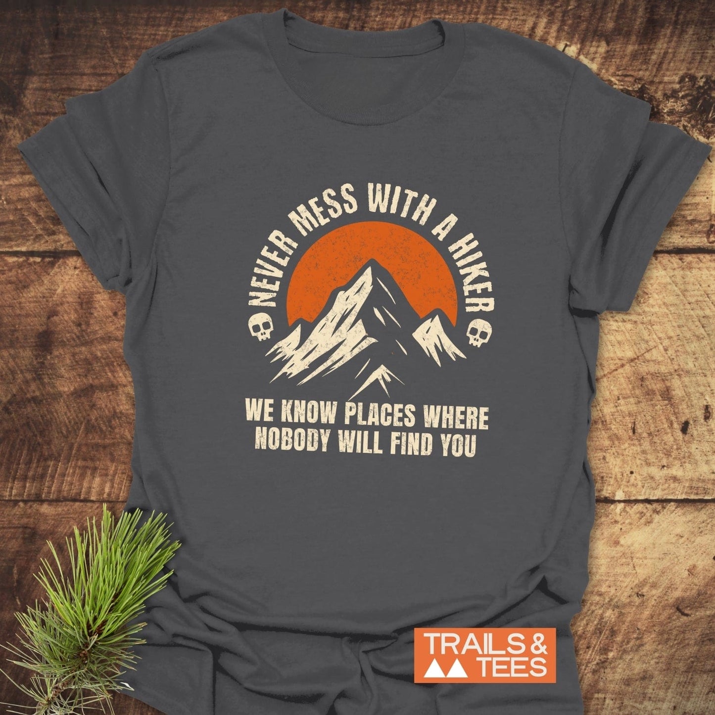 Never Mess With A Hiker T-Shirt featuring a mountain graphic with an orange sun and skulls, laid on wood next to a pine sprig.