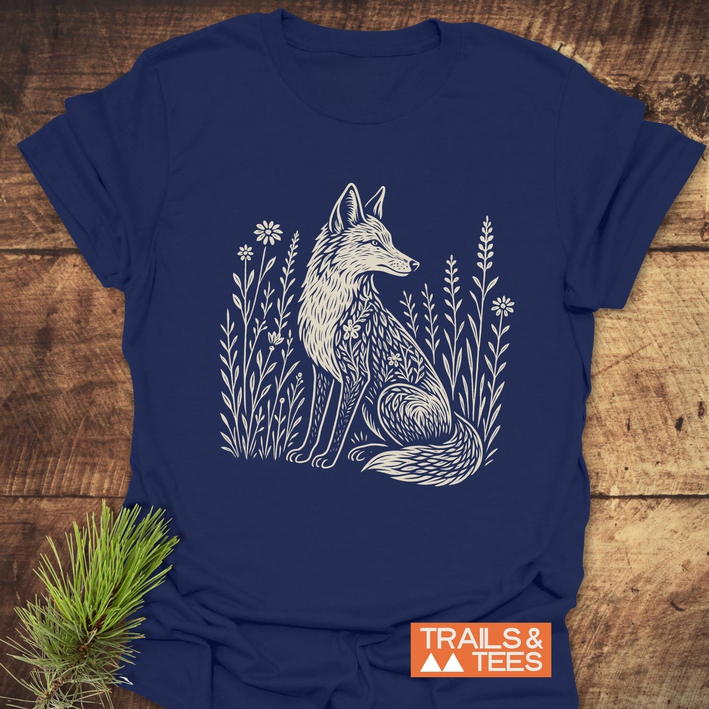 Wildflower Fox T-Shirt featuring a stylized fox among wildflowers, printed in off-white on navy-blue, laid on rustic wood with a pine sprig accent.