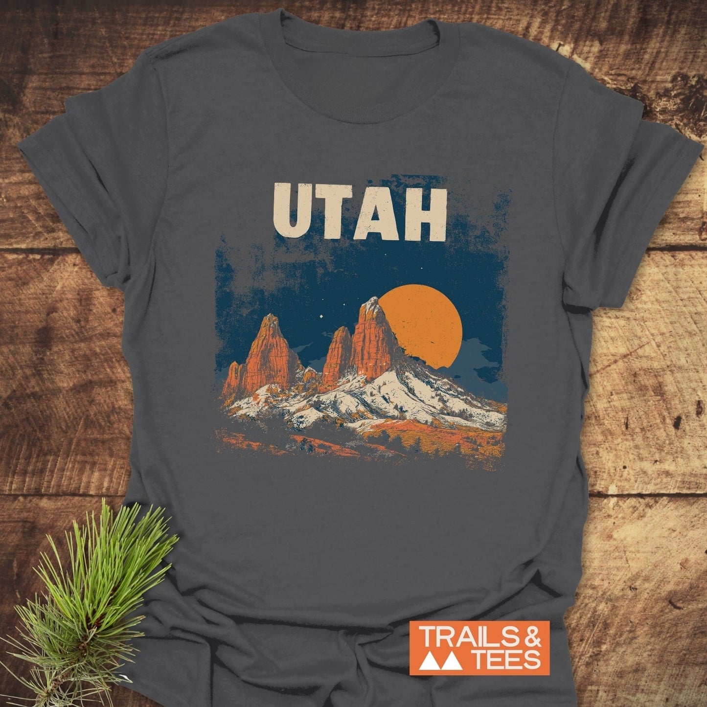 Utah Retro T-Shirt with mountain scene graphic, folded on wood surface with pine sprig. Made from soft cotton, ideal for outdoor adventures.