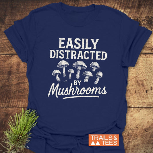 Easily Distracted By Mushrooms T-Shirt features a white mushroom design on a navy tee, laid flat with TRAILS & TEES logo and pine sprig.