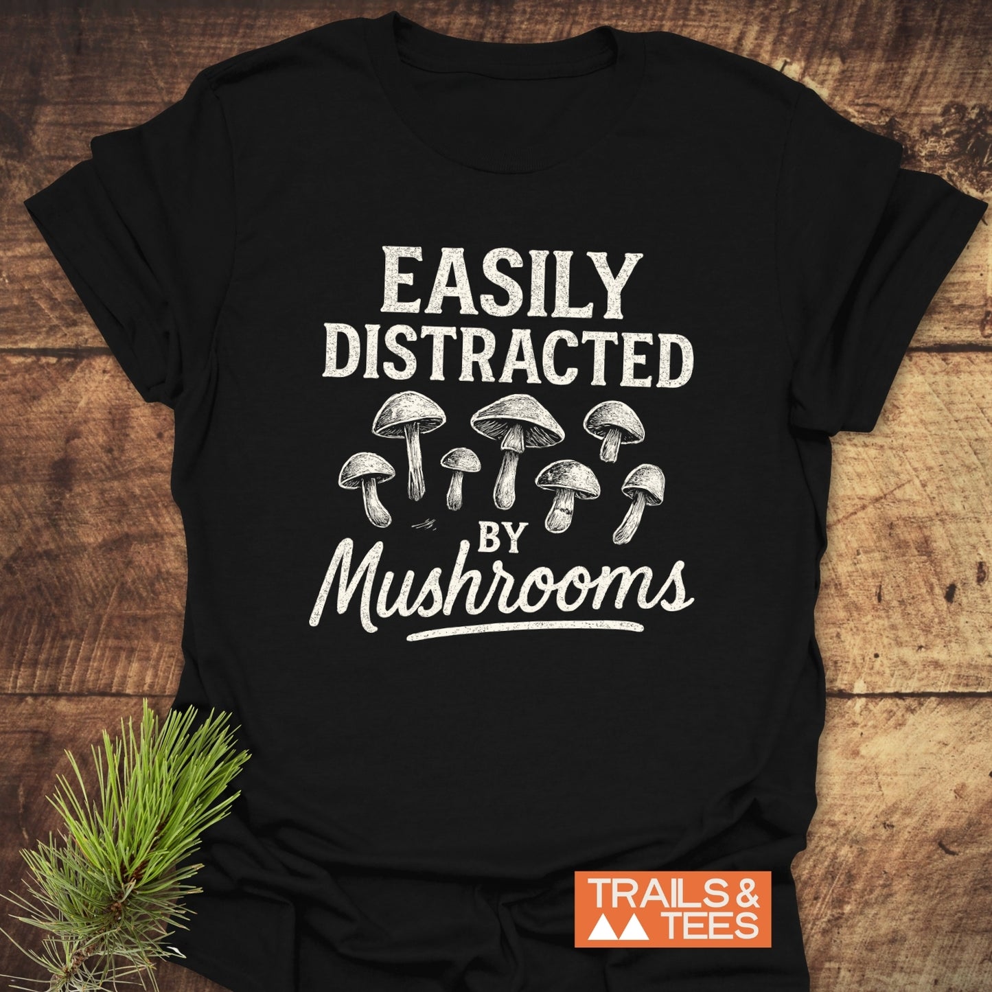 Black T-shirt with “Easily Distracted by Mushrooms” in white, featuring line-drawn mushrooms. Logo “TRAILS & TEES.” visible. Made from soft, lightweight cotton.