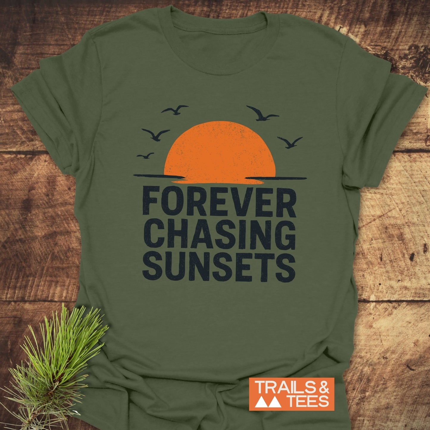 Forever Chasing Sunsets T-Shirt with sunset print and seabirds, featuring soft ring-spun cotton for comfort. Includes Trails & Tees logo. Laid on rustic wood with pine sprig.