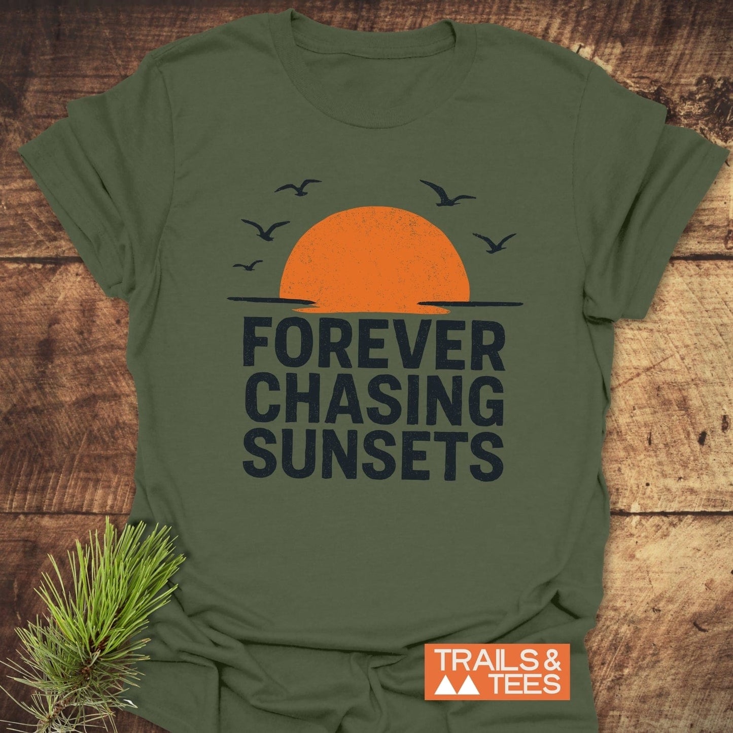 Forever Chasing Sunsets T-Shirt with sunset print and seabirds, featuring soft ring-spun cotton for comfort. Includes Trails & Tees logo. Laid on rustic wood with pine sprig.