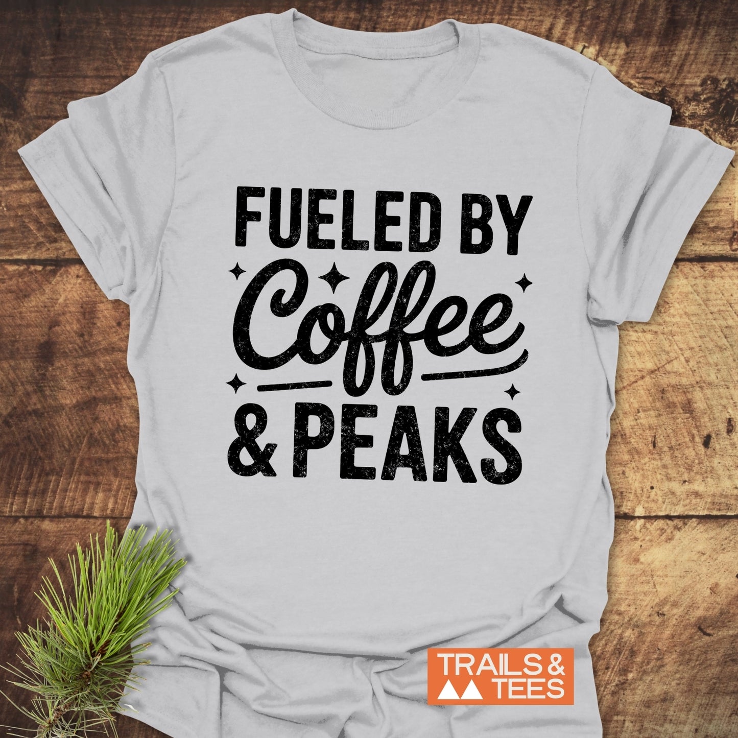 Fueled By Coffee And Peaks T-Shirt on rustic wood, featuring black lettering. Soft, lightweight cotton, pre-shrunk with durable print, ideal for outdoor enthusiasts.