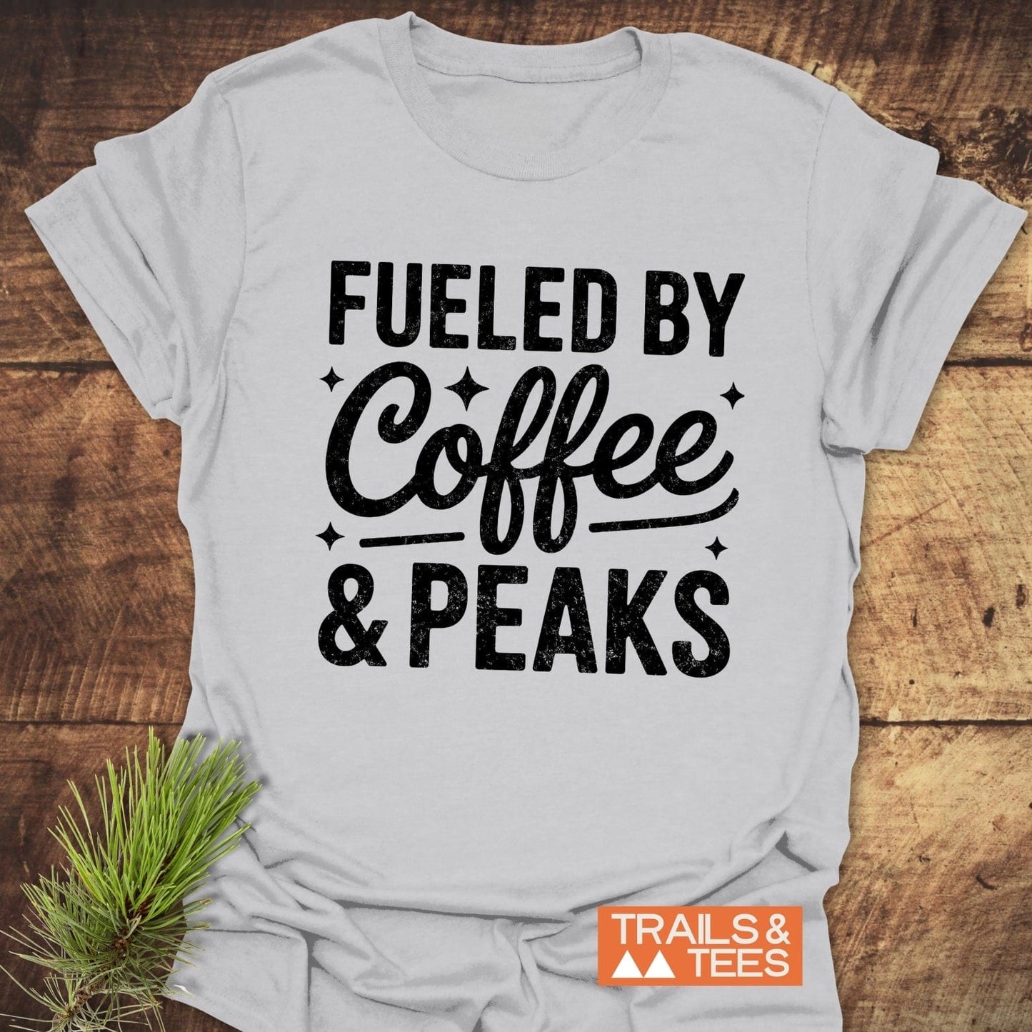Fueled By Coffee And Peaks T-Shirt on rustic wood, featuring black lettering. Soft, lightweight cotton, pre-shrunk with durable print, ideal for outdoor enthusiasts.