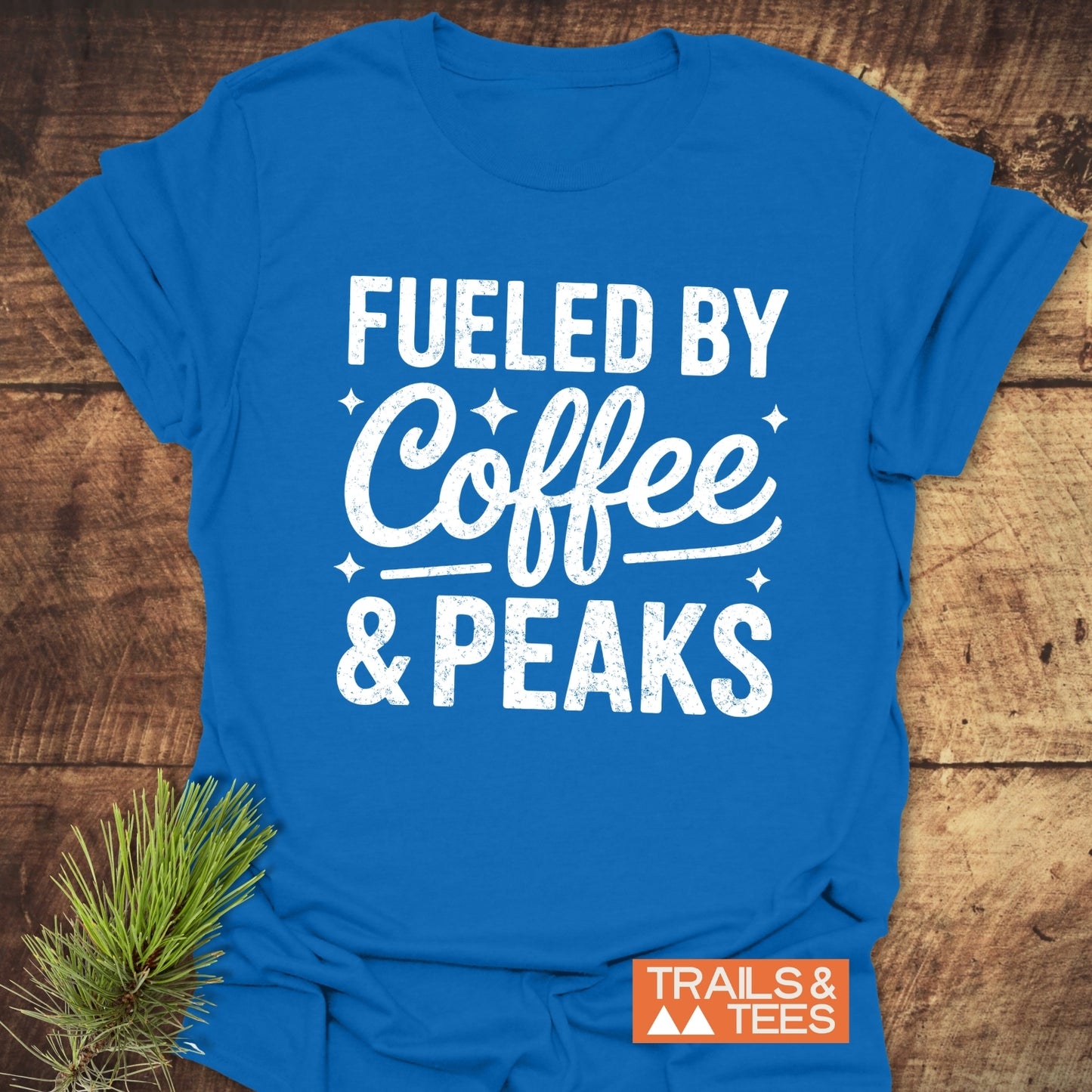 Flat-lay of the Fueled By Coffee And Peaks T-Shirt, featuring a bold white graphic and small logo, laid on wood with a pine sprig accent.