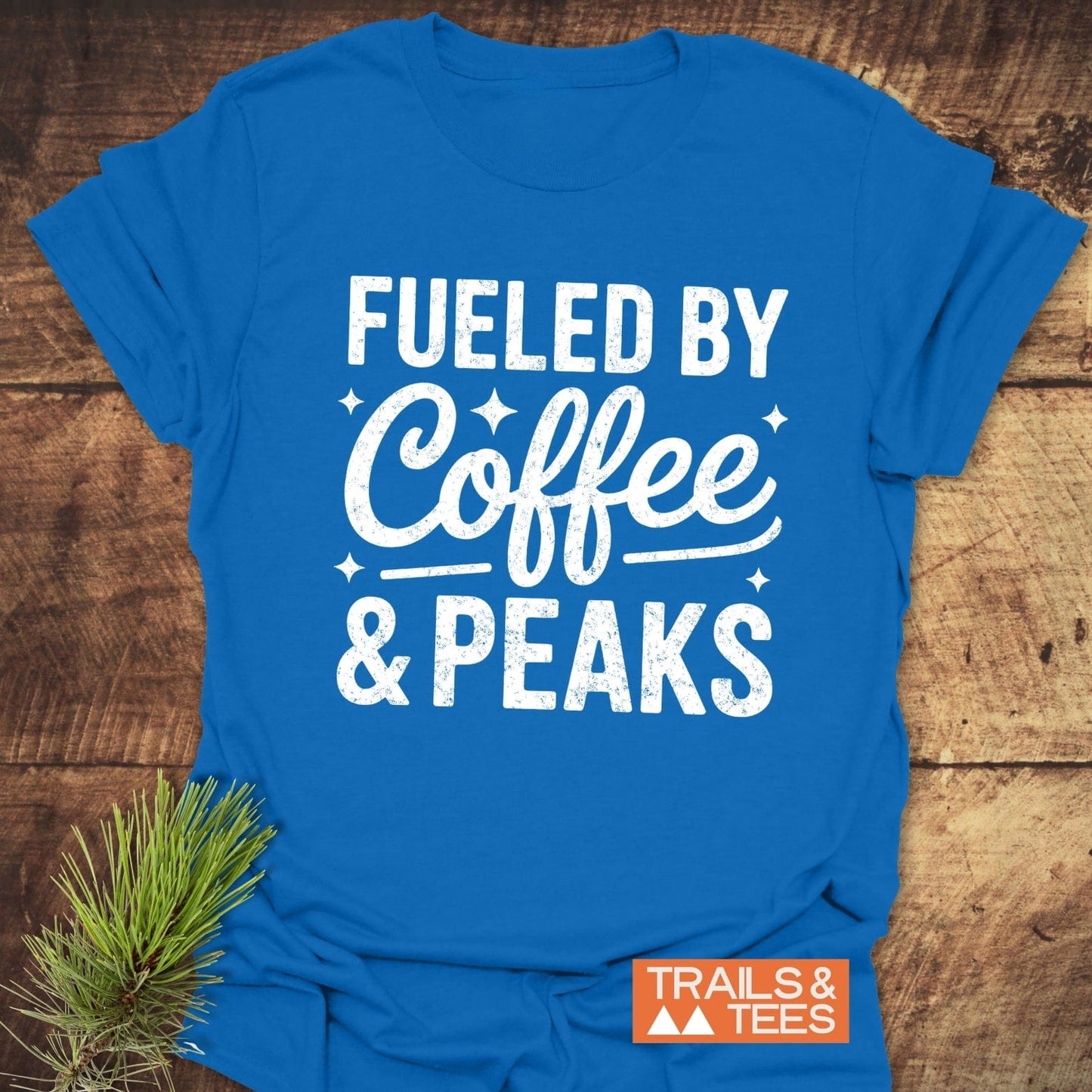 Flat-lay of the Fueled By Coffee And Peaks T-Shirt, featuring a bold white graphic and small logo, laid on wood with a pine sprig accent.