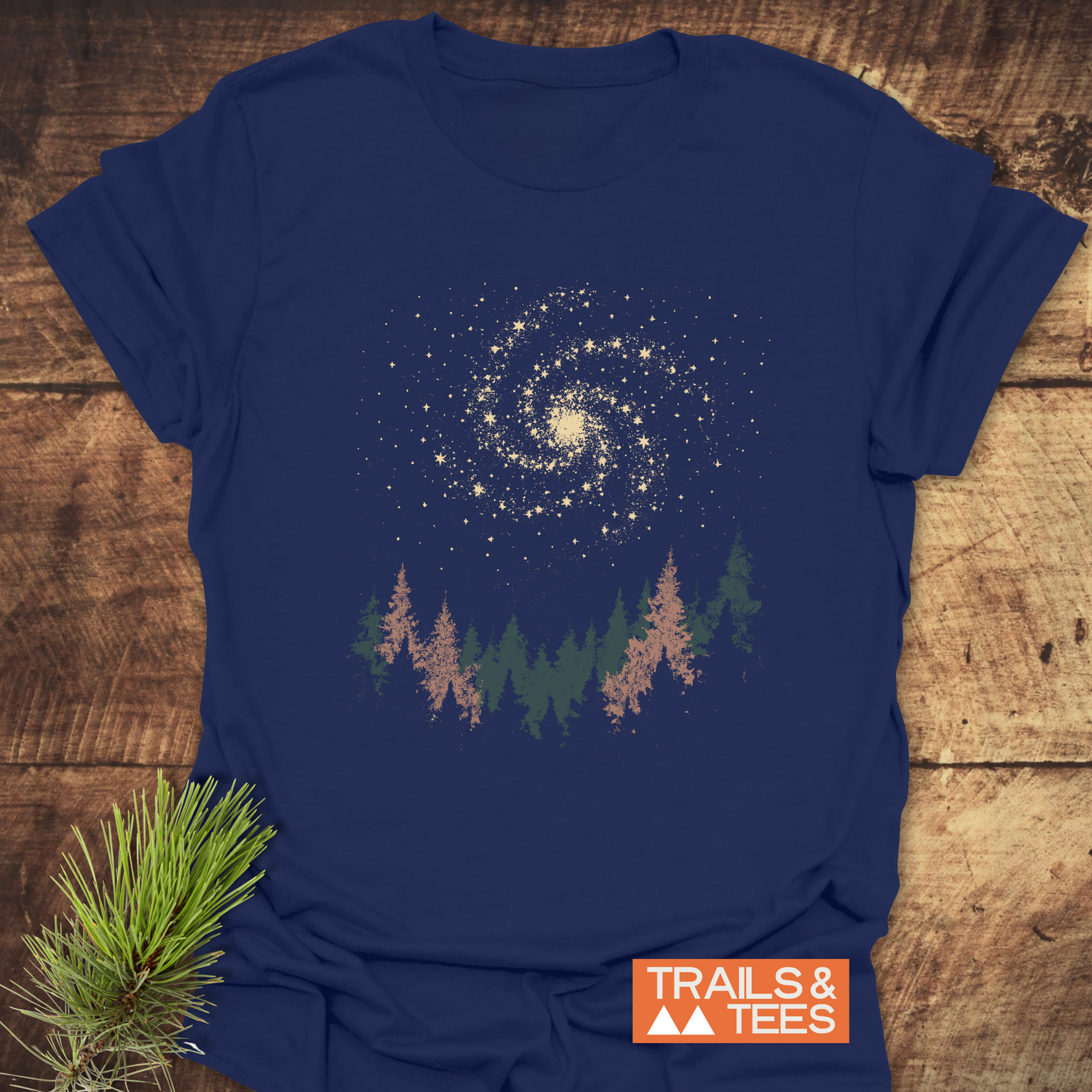 Woodland Nightsky T-Shirt featuring a forest treeline and galaxy design, laid flat on wood, showcases soft, durable, pre-shrunk cotton for everyday adventure.