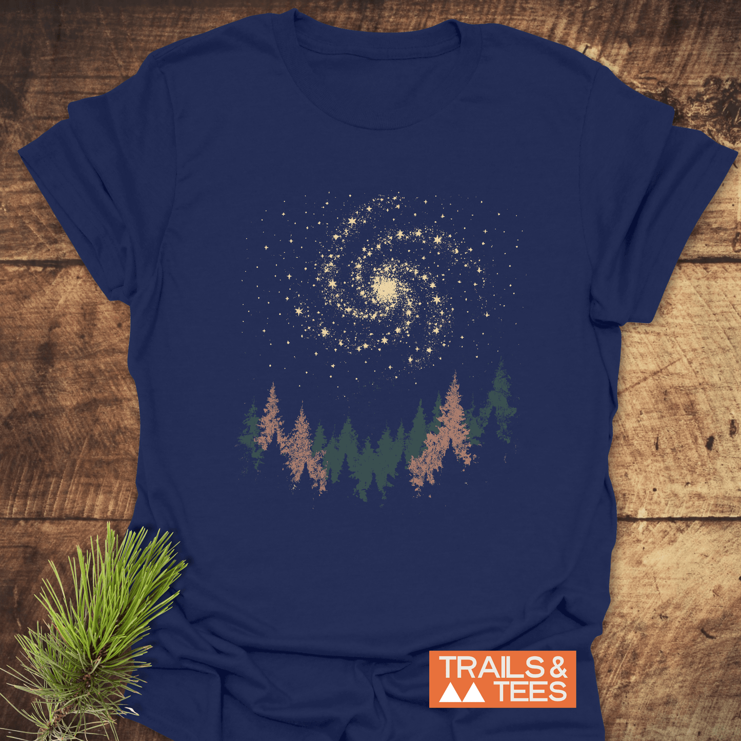 Woodland Nightsky T-Shirt featuring a forest treeline and galaxy design, laid flat on wood, showcases soft, durable, pre-shrunk cotton for everyday adventure.