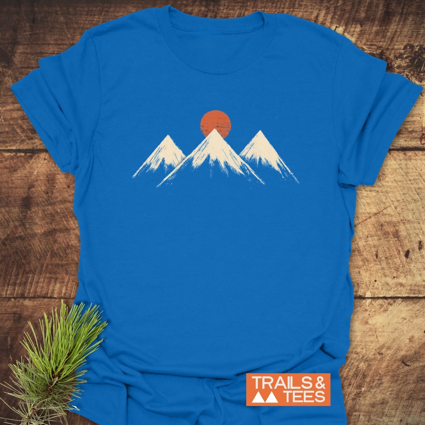 Three Mountains And Sun T-Shirt featuring snow-capped peaks and sun graphic, laid on wood. Crafted from soft cotton, perfect for outdoor enthusiasts.